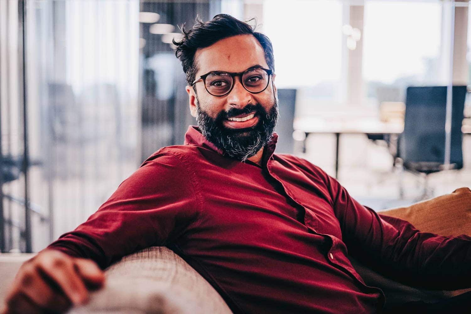 A smiling Rahman sitting on a couch looks into the camera.